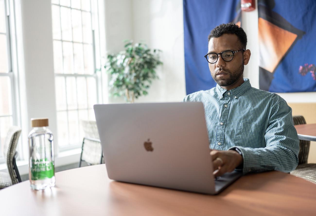 A man sitting at a desk in front of a window using a laptop