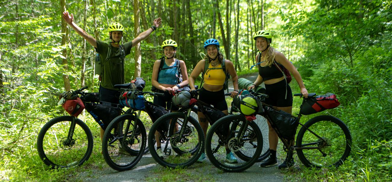 Group of four students standing with their mountain bikes on a trail in the woods.
