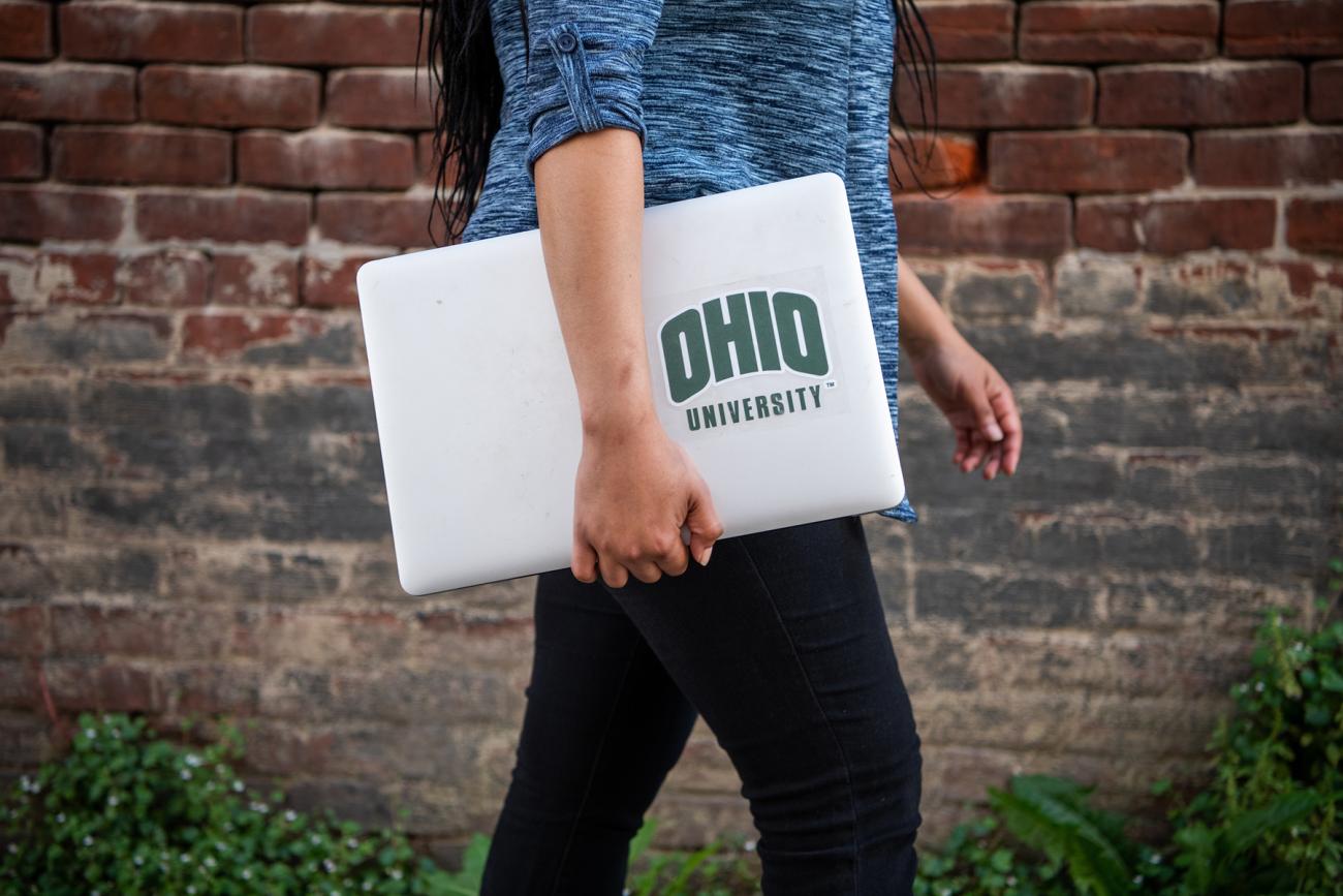 Person walking with a laptop under their arm with an OHIO University sticker on it.
