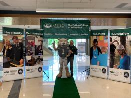 Curt Plummer stands on a green carpet to welcome visitors to Patton Hall surrounded by department banners