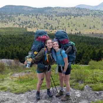 Two education students pose and smile while wearing hiking packs in the mountains