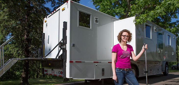 Person standing in front of a trailer with windows, a small room protruding from the side, and a platform and stairs attached to the rear door