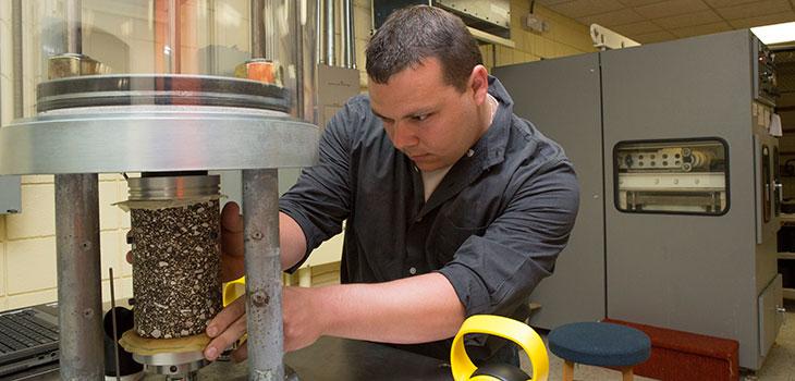 Person in lab adjusting a cylindrical piece of asphalt held in a machine
