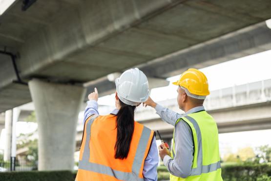 two workers point at and inspect a bridge while underneath it.