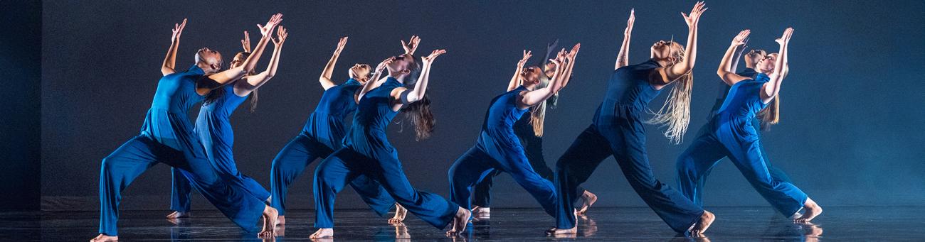 A group of students dressed in dark blue hold a pose during a dance performance in front of a dark blue background.
