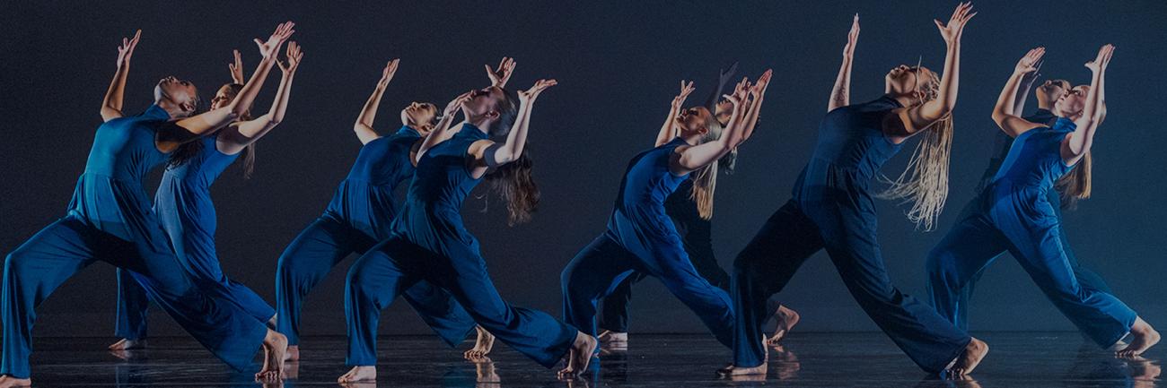 A group of students dressed in dark blue hold a pose during a dance performance in front of a dark blue background.