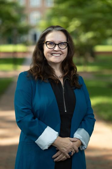Dean Schroeder-Arce posing for a portrait on college green, wearing black-framed glasses, a black shirt, and a deep teal blazer