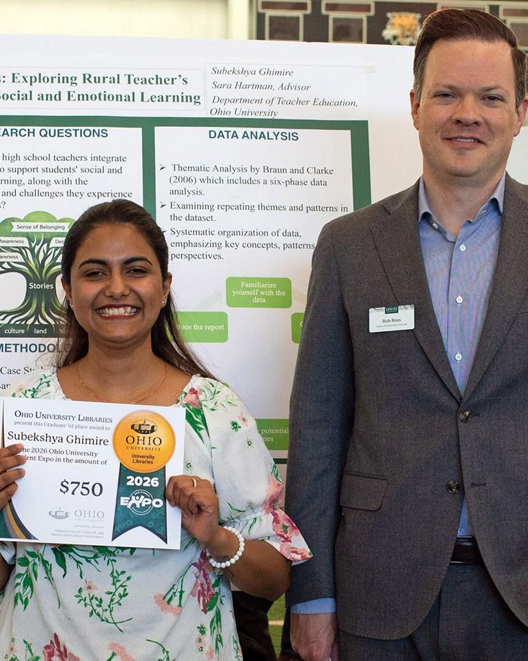 Person stands holding award certificate in front of their research poster. Tall man in grey suit stands next to them.