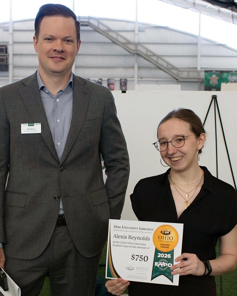 Person stands holding award certificate in front of their research poster. Tall man in grey suit stands next to them.