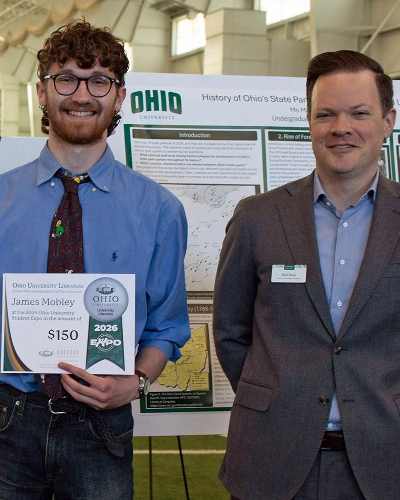 Person stands holding award certificate in front of their research poster. Tall man in grey suit stands next to them.