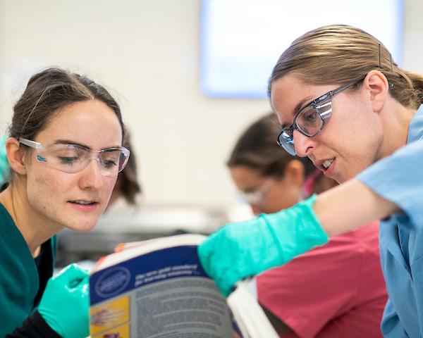 A student looks at a textbook with an instructor in the anatomy lab