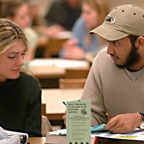 Two students seated at a table reading a book. 