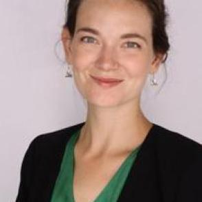 A white woman with dark hair pulled back smiling facing the screen in a dark green blouse and black blazer