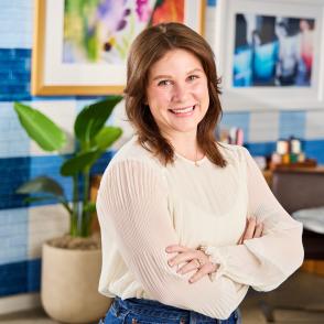 A headshot of a woman with shoulder length brown hair smiling facing the camera wearing a white blouse. She is standing in a bright room with white and blue stripped wallpaper. 