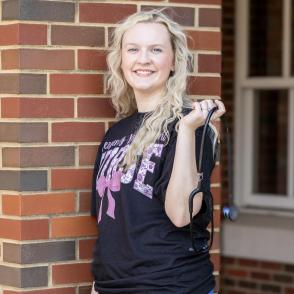 Woman wearing a shirt that reads 'nurse' smiling against brick background