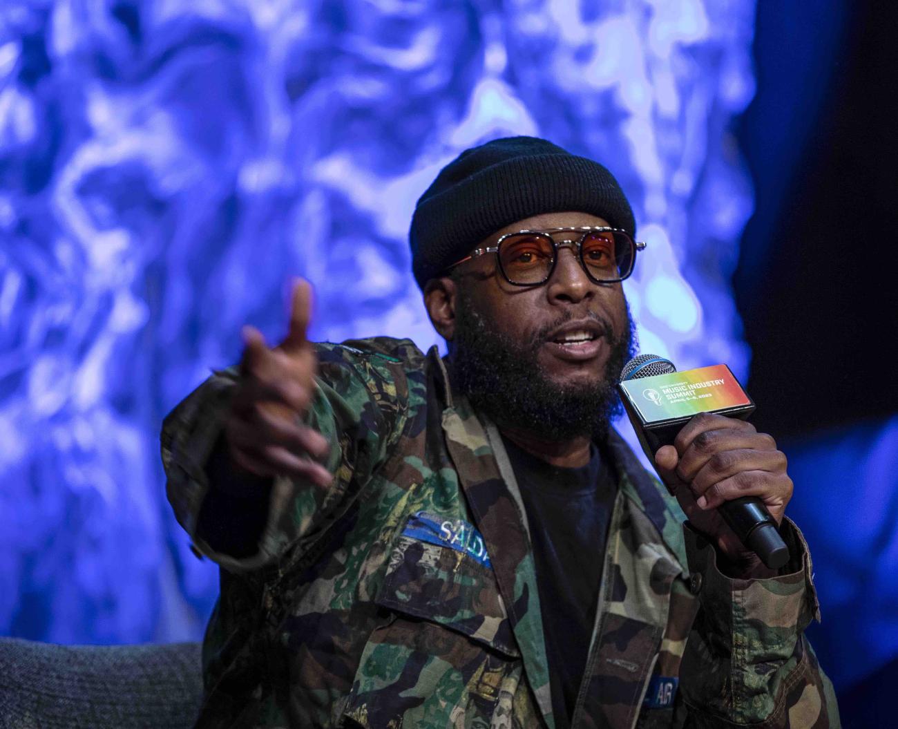 Talib Kweli speaking into microphone while extending right arm forward