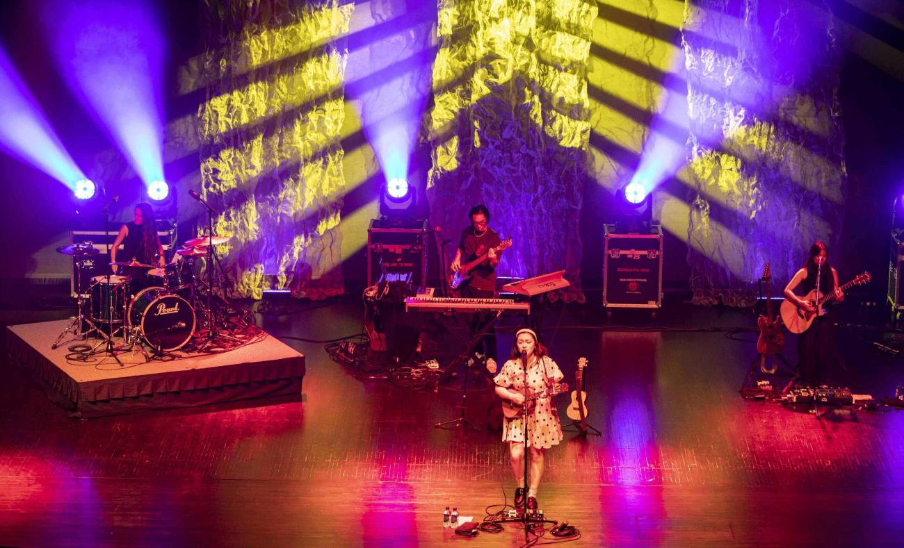 mxmtoon singing on microphone with bandmates in background