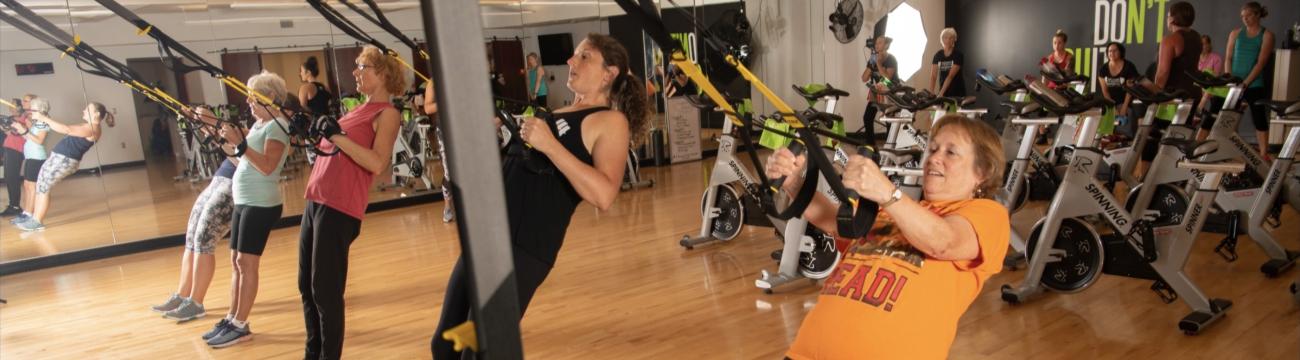 WellWorks members in group fitness room using TRX trainers and standing near spin bikes.