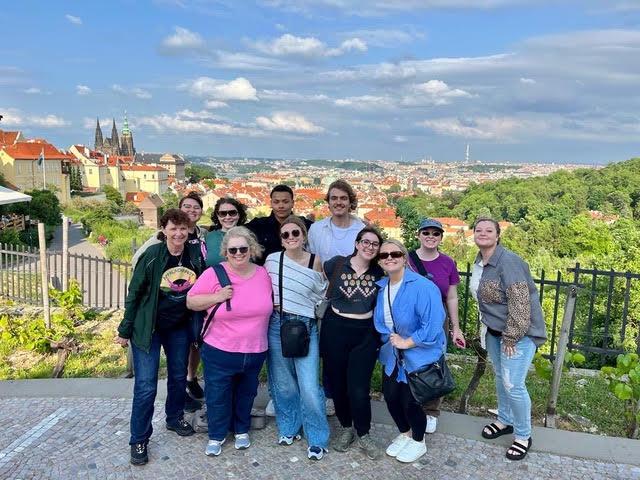 A group of 11 pose for a photo against a European city backdrop. The wind is blowing.
