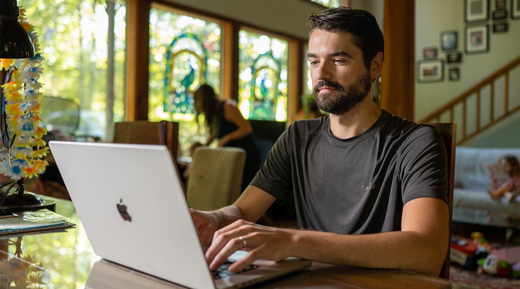 A man does work on his laptop while his family mills about the living room behind him.