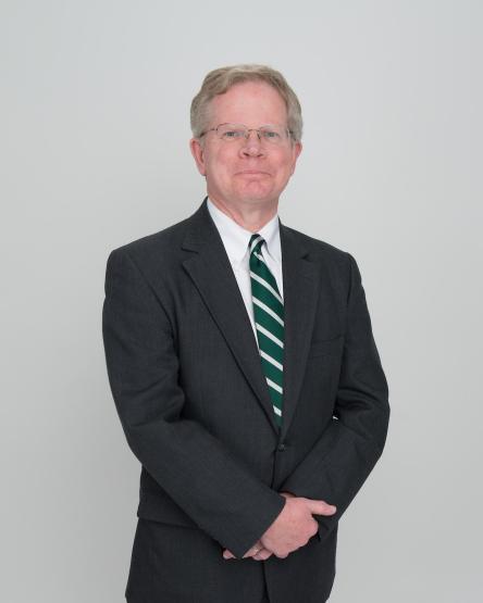 Dr. Richard W. Greenlee, wearing a black suit, with a green and white tie, standing with his hands together