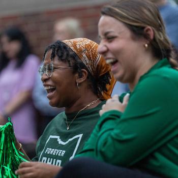 two female students laughing on college green in green Ohio sweatshirts 